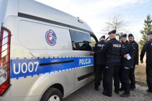 Grupa umundurowanych policjantów stojących przy otwartych prawych drzwiach radiowozu. Na radiowozie umieszczony numer boczny 007 i  napis Policja oraz logo z twarzą rzymskiego żołnierza i napisem Praetorian