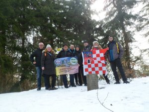 Zdjęcie Kolorowe. Grupa ośmiu osób, w zimowej scenerii pozuje do zdjęcia w górach trzymając flagę Kalisza oraz Koła PTTK „Z Buta”.