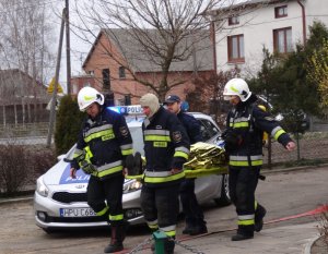 Zdjęcie kolorowe. Trzech strażaków i policjant niosą deskę ortopedyczną, na której transportują osobę. W tle oznakowany radiowóz Policji.