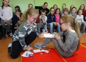 Zdjęcie kolorowe. Na środku klasy na materacu siedzi dziewczynka, której koleżanka bandażuje prawą rękę. Za nimi w tle siedzą uczniowie, którzy przyglądają się scence.