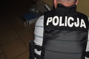 Zdjęcie kolorowe. Policjant operacyjny Wydziału do walki z Przestępczością Gospodarczą, stojący tyłem ubrany w kamizelkę taktyczną z napisem &quot;POLICJA&quot;, liczy zabezpieczone paczki papierosów.