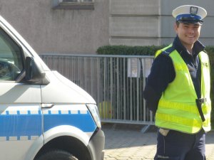 Zdjęcie kolorowe. Policjant ruchu drogowego w umundurowaniu służbowym i kamizelce odblaskowej, stojący obok oznakowanego radiowozu.