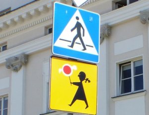 Zdjęcie kolorowe. Zdjęcie znaków drogowych zamontowanych na jednym słupku. U góry znak drogowy D-6 &quot;przejście dla pieszych&quot;, u dołu T-27 &quot;tabliczka wskazująca, że przejście dla pieszych jest szczególnie uczęszczane przez dzieci&quot;.
