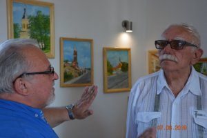 Zdjęcie kolorowe. Marian Ozdowski podczas rozmowy z jedną z osób odwiedzających wernisaż.  W tle obrazy na ścianach.