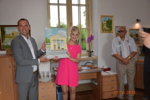Zdjęcie kolorowe. Mężczyzn i kobieta trzymający obraz Mariana Ozdowskiego na którym namalowany jest budynek Rogatki Kaliskiej.