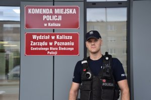 Zdjęcie kolorowe. Policjant w umundurowaniu służbowym na tle fasady Komendy Miejskiej Policji w Kaliszu.