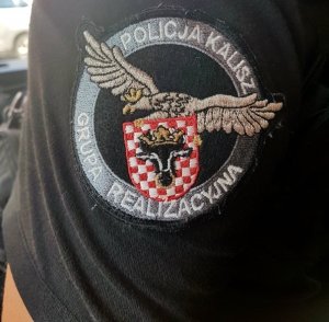 Zdjęcie kolorowe. Policjant ubrany w czarny t-shirt z naszywką na ramieniu &quot;Policja Kalisz. Grupa Realizacyjna&quot;