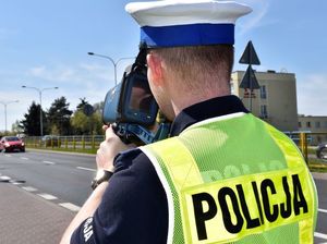 Zdjęcie kolorowe , na nim policjant ruchu drogowego trzymający w ręku przyrząd służący do pomiaru prędkości